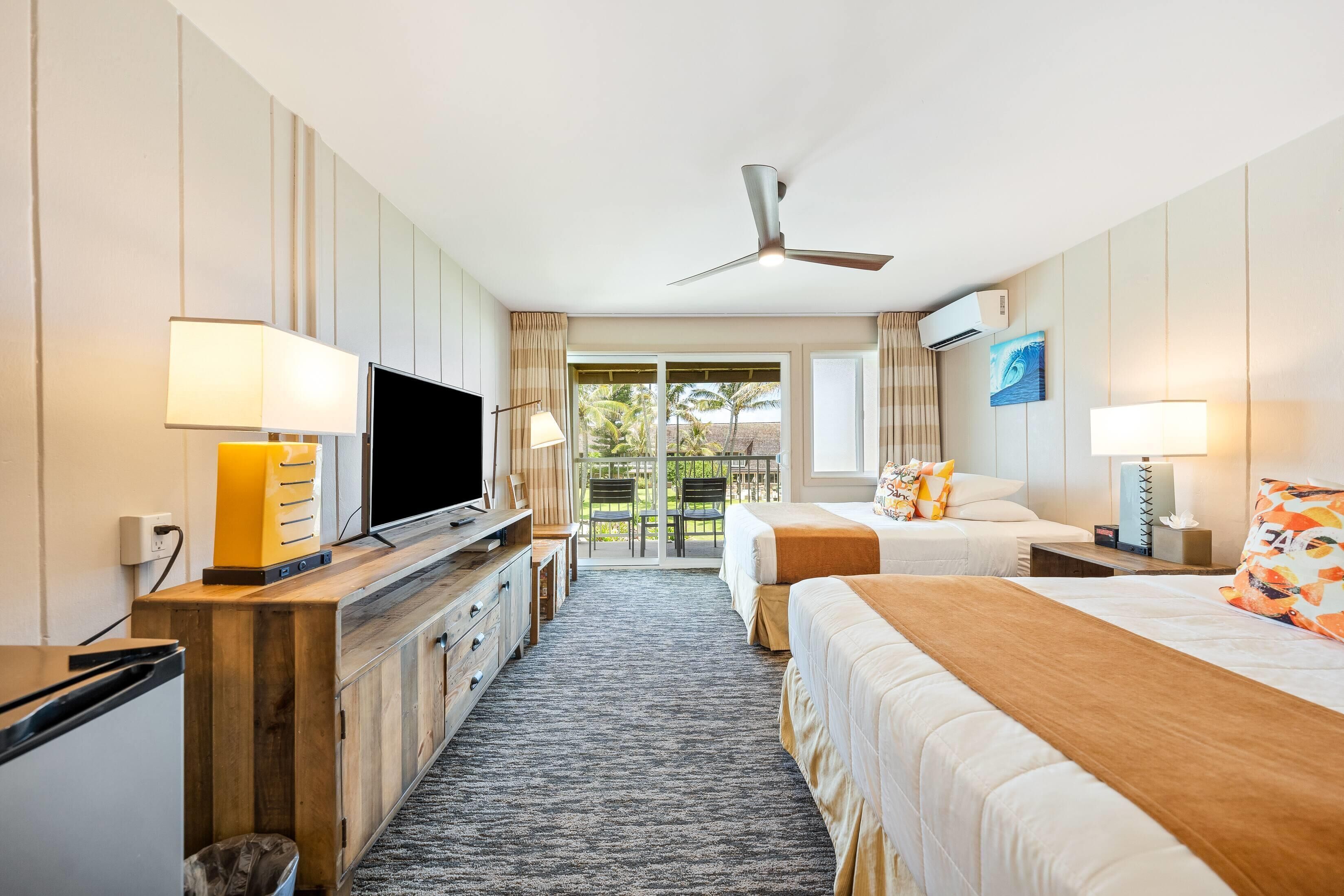A hotel room with a TV, wooden cabinet, two beds, a ceiling fan, and a balcony with a view of palm trees.