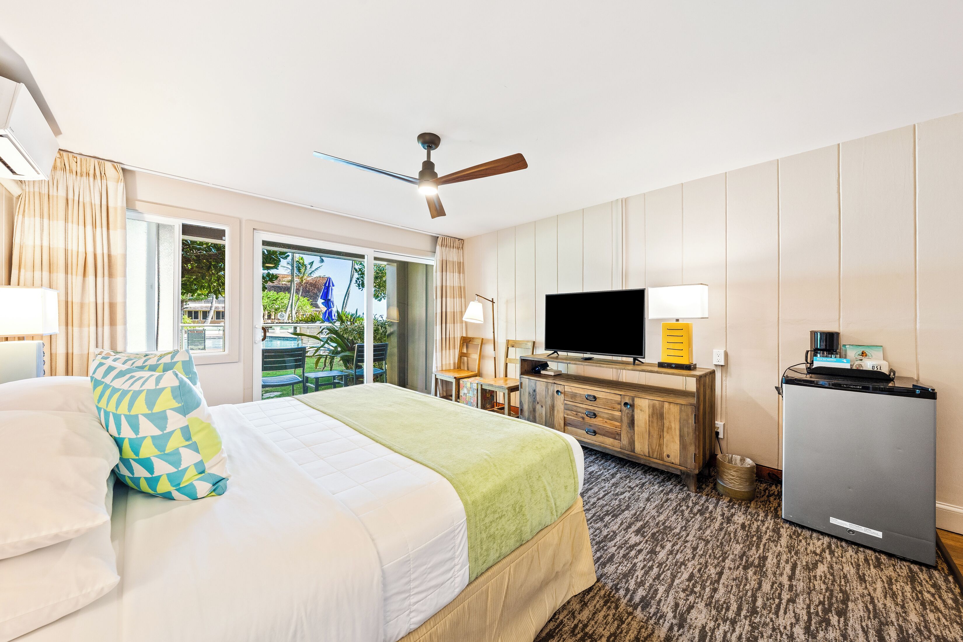 A hotel room with a bed, a desk with a television, a ceiling fan, and a view of the outdoors.