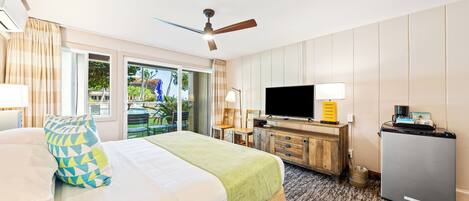 A hotel room with a bed, a desk with a television, a ceiling fan, and a view of the outdoors.