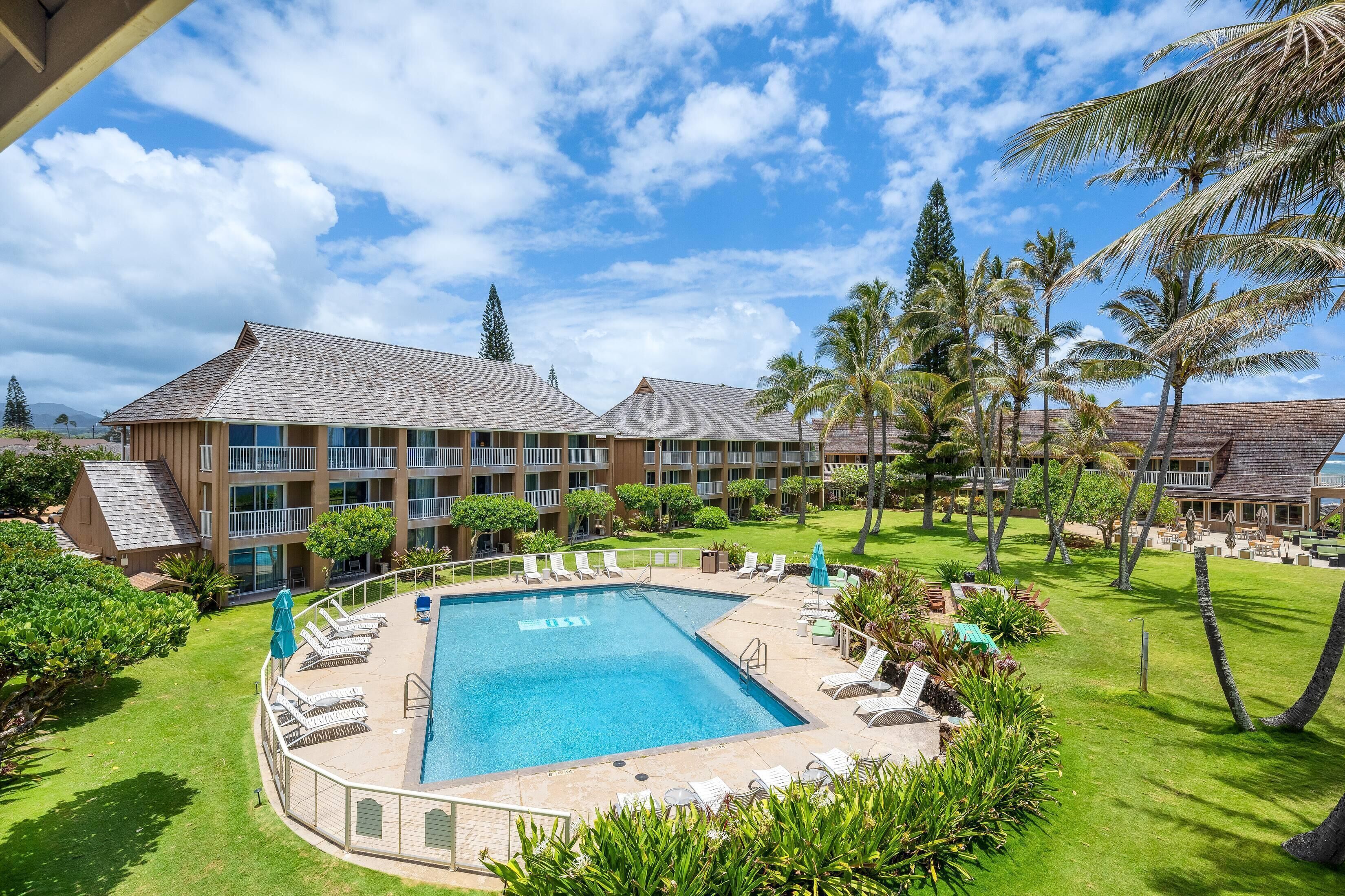 A hotel with a swimming pool surrounded by lounge chairs, palm trees, and a well-maintained lawn.
