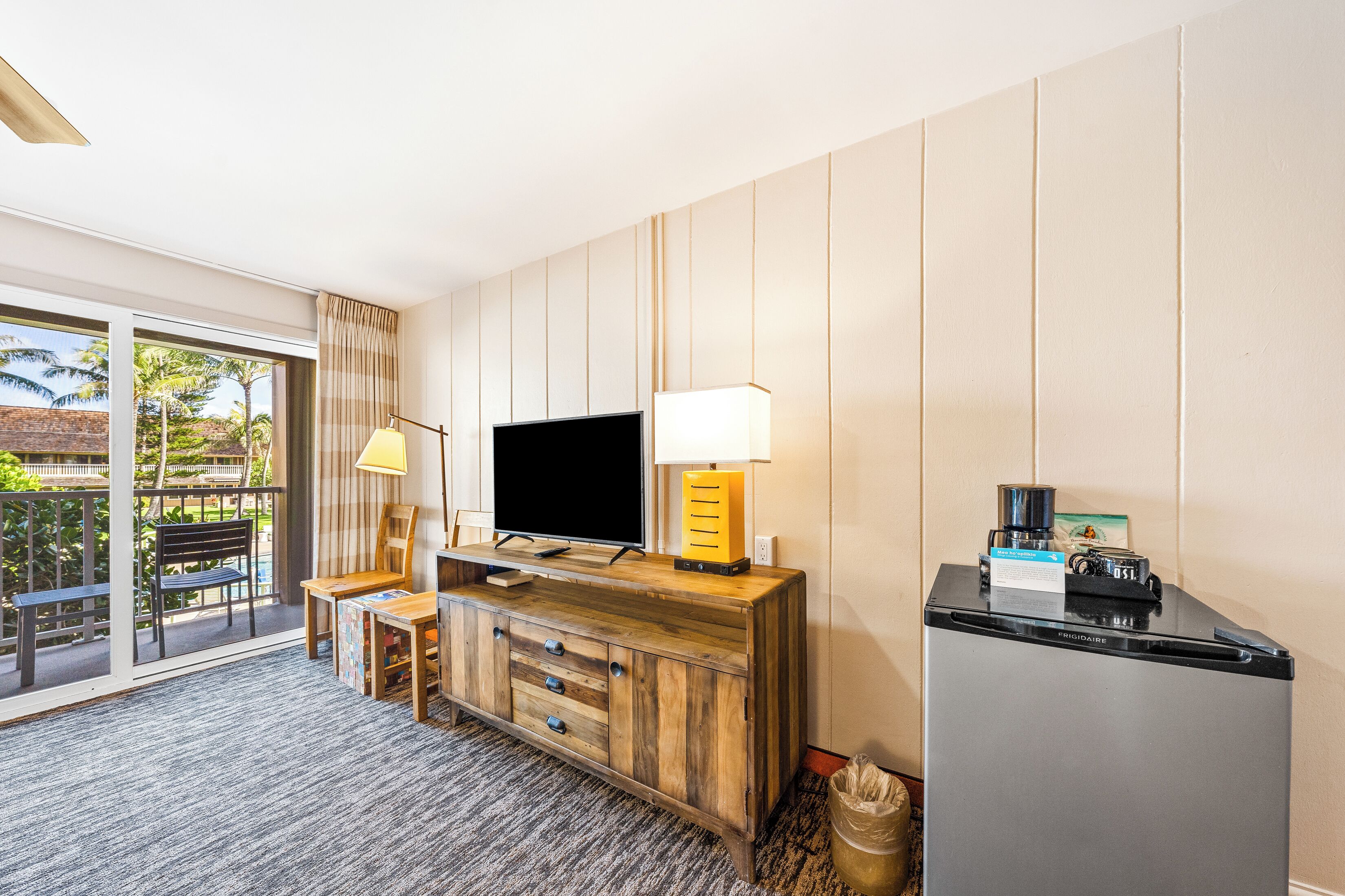 A hotel room with a wooden TV stand, a flat-screen TV, a lamp, a refrigerator, and a view of the outdoors.