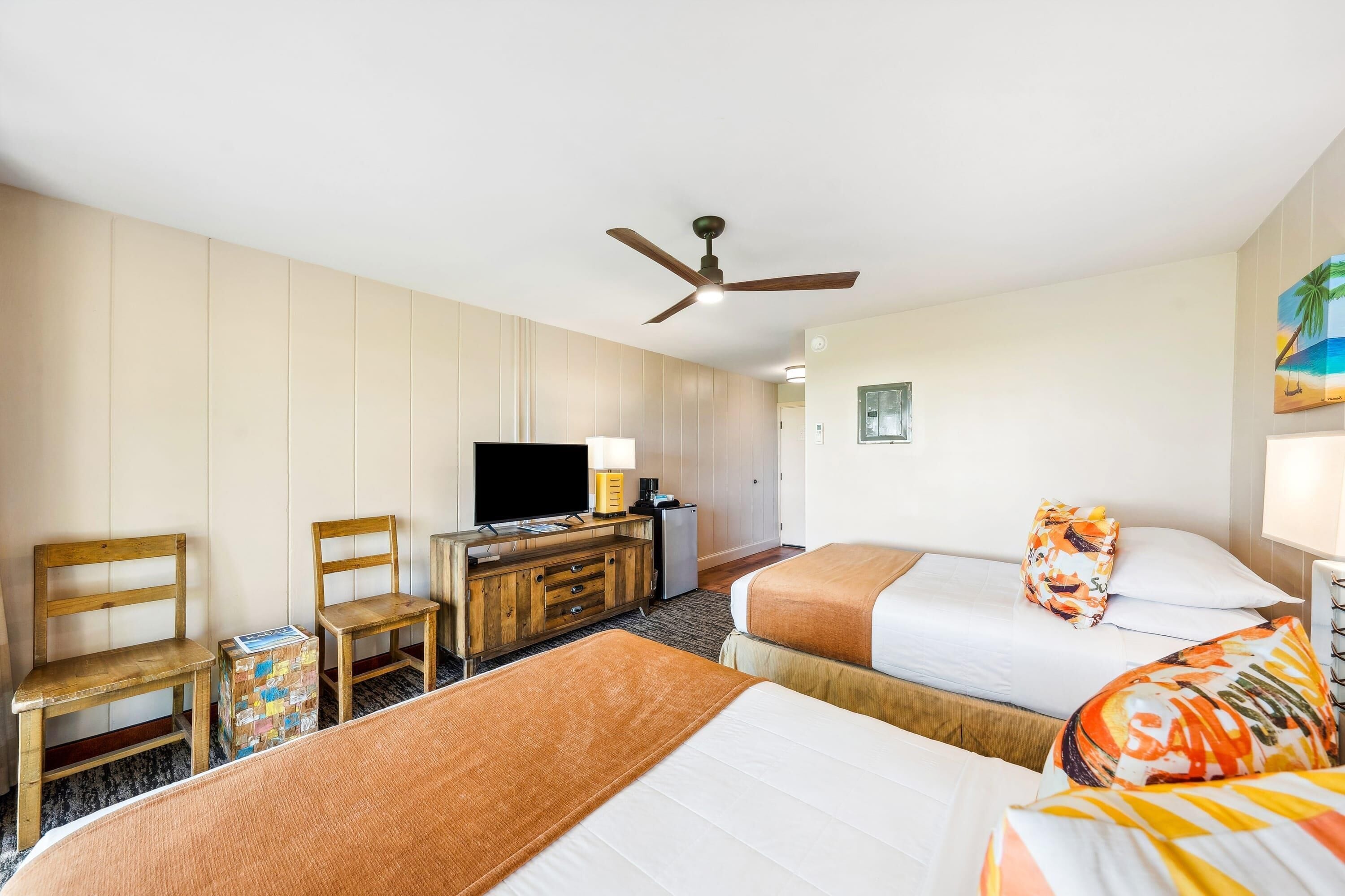 A hotel room with two beds, a television, a desk with a lamp, and a ceiling fan.