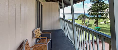 Un balcón techado con dos sillas de madera y una mesa, con vistas a una zona verde con árboles y el océano.