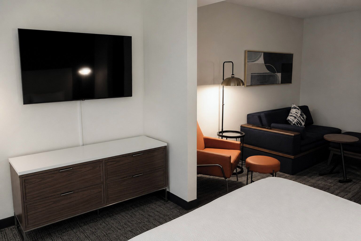 Habitación de hotel con televisión de pantalla plana en la pared, sofá de color oscuro, sillón naranja, una mesita redonda y una lámpara de pie.