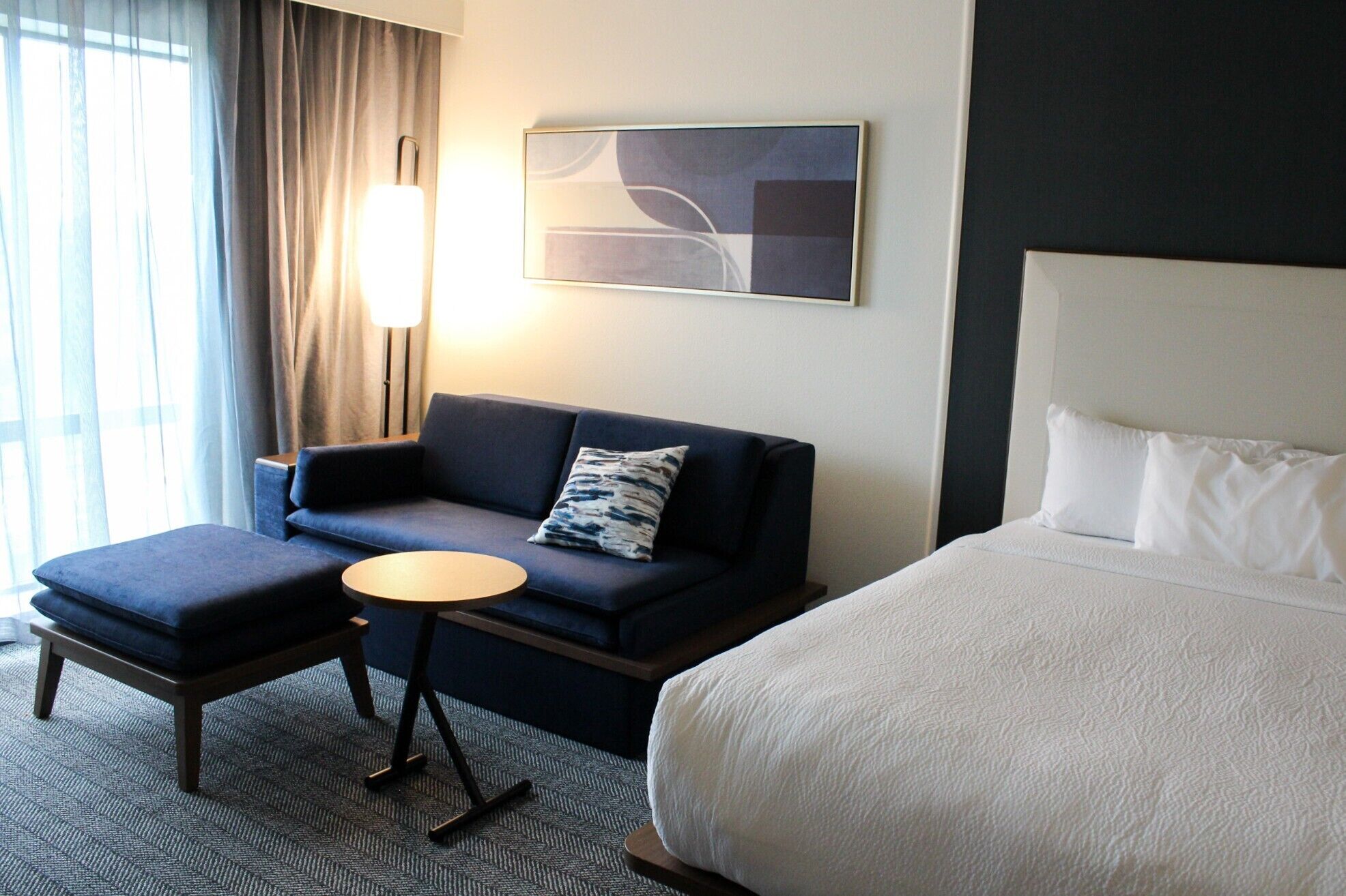 A hotel room with a bed, a sofa, a small table, and a lamp.