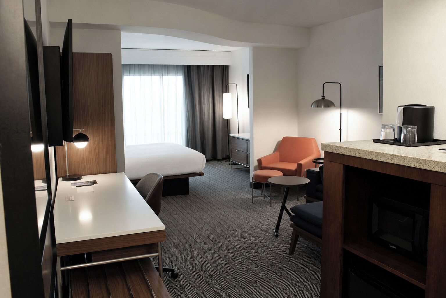 Ett hotellrum med en säng, ett skrivbord, en stol, ett litet bord, en kaffemaskin och en tv.