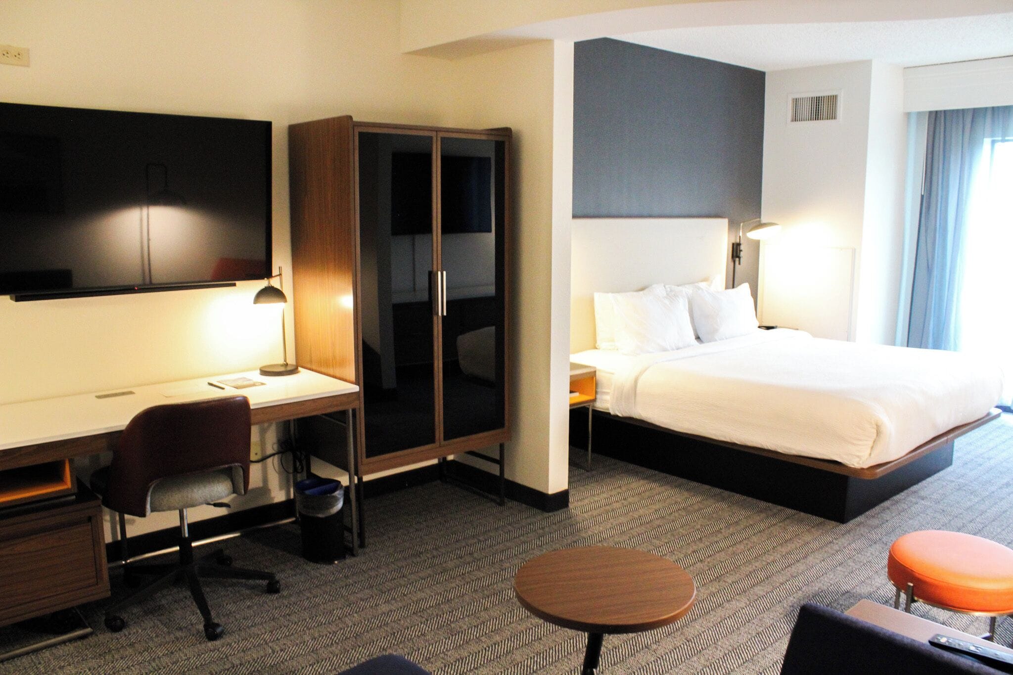 Une chambre d’hôtel équipée d  ’un bureau, d’une chaise, d’une armoire, d’un lit, d’une table de chevet, d’une lampe et d’une petite table ronde.