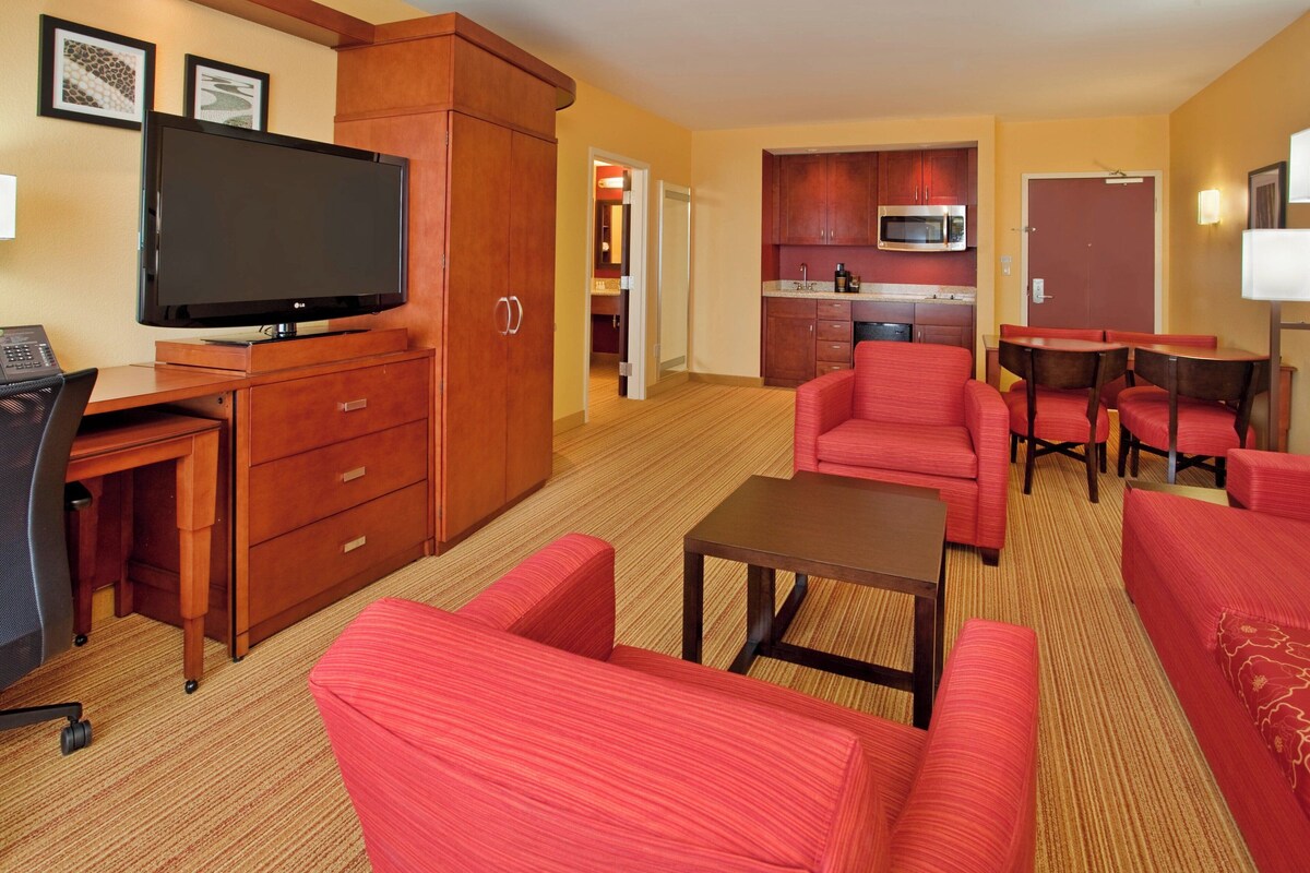 Ein Hotelzimmer mit Fernseher, Schreibtisch, Küchenzeile und roten Polstermöbeln.