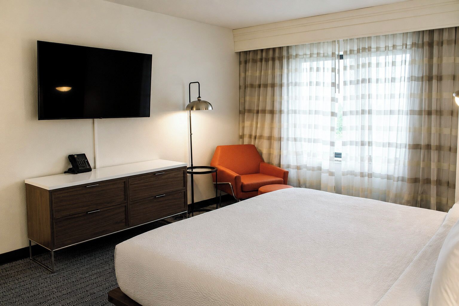 Habitación de hotel con cama, escritorio con teléfono, silla, televisor montado en la pared y ventana con cortinas.