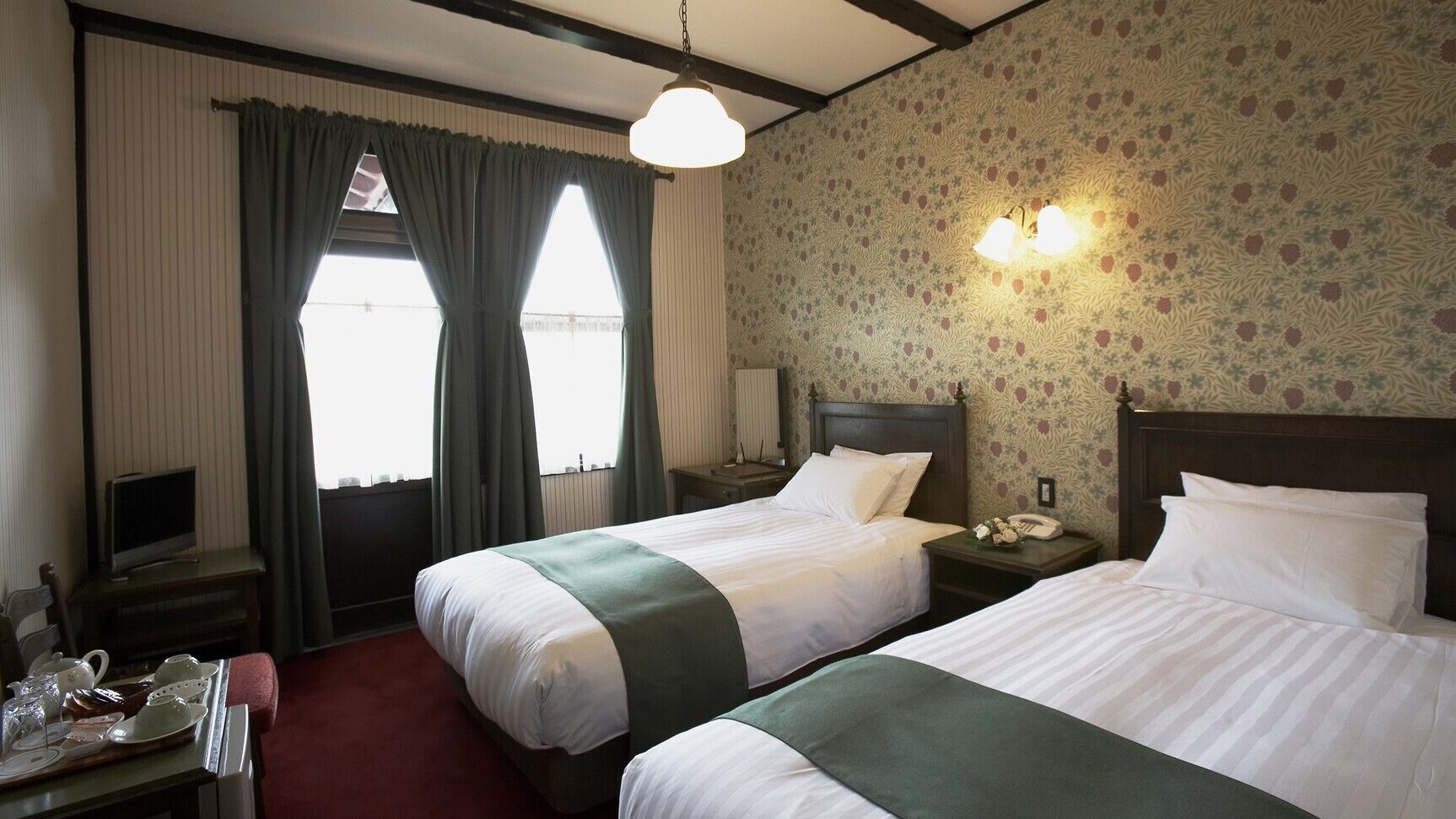 A hotel room with two beds, a television, a desk with a telephone, and a window with curtains.