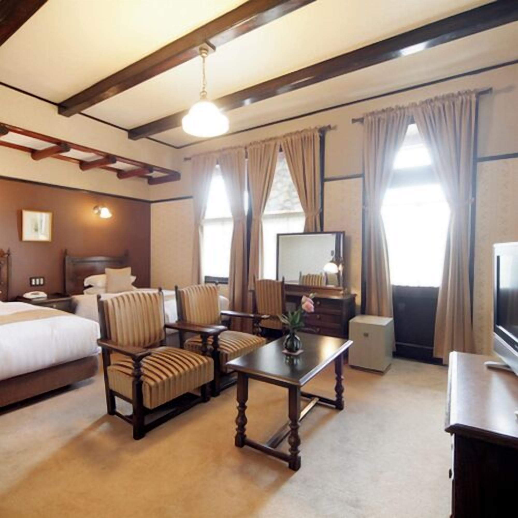 A hotel room with a bed, two armchairs, a small table with a vase, a television, and a dresser.
