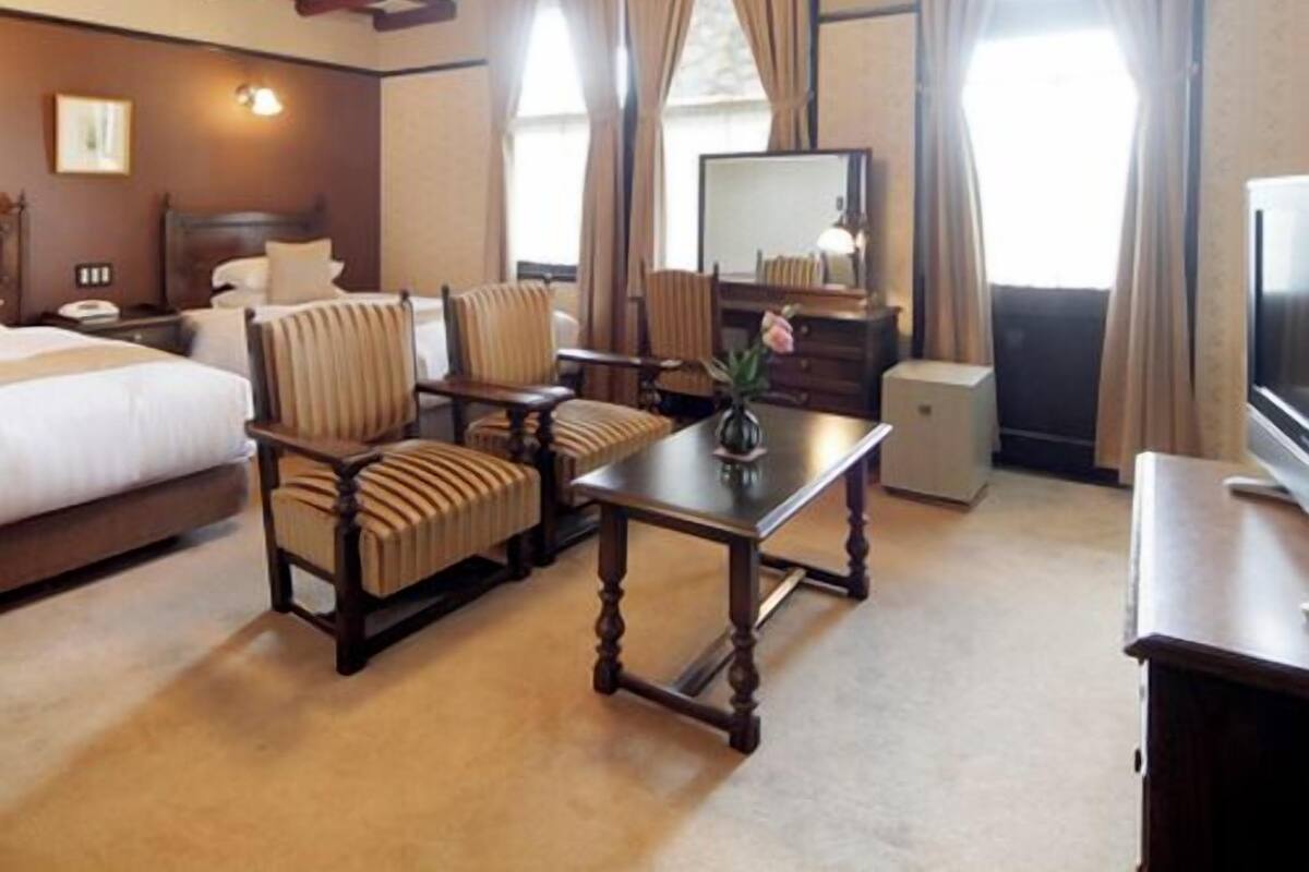 A hotel room with a bed, two armchairs, a small table with a vase, a television, and a dresser.