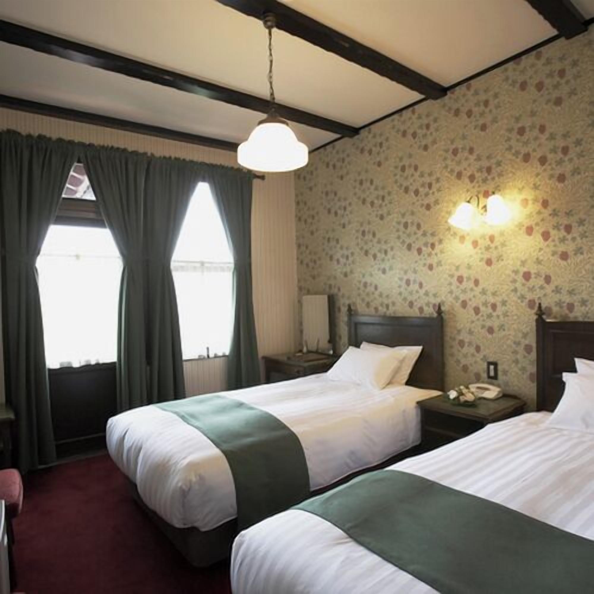 A hotel room with two beds, a window with curtains, a floral wallpaper, and a nightstand with a lamp.