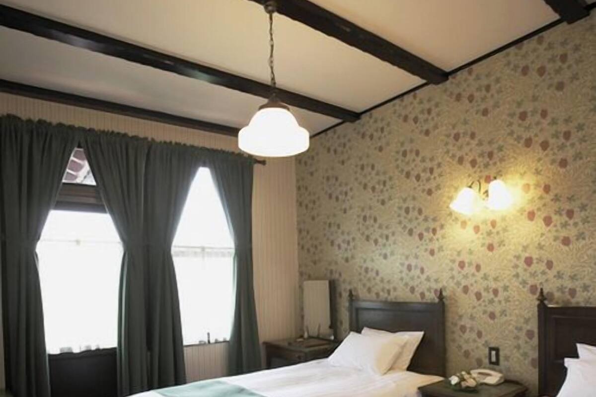 A hotel room with two beds, a window with curtains, a floral wallpaper, and a nightstand with a lamp.