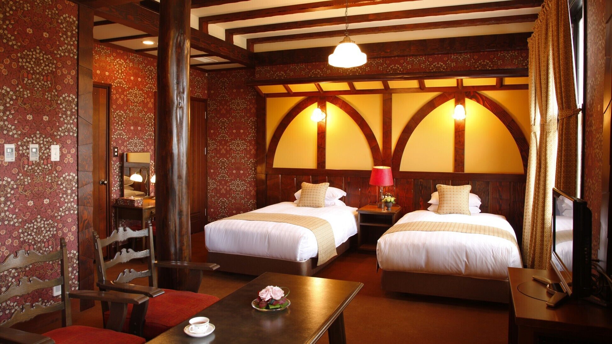 A hotel room with two beds, a wooden chair, a table with a cup and flowers, and a TV.