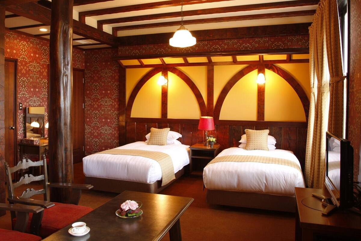 A hotel room with two beds, a wooden chair, a table with a cup and flowers, and a TV.