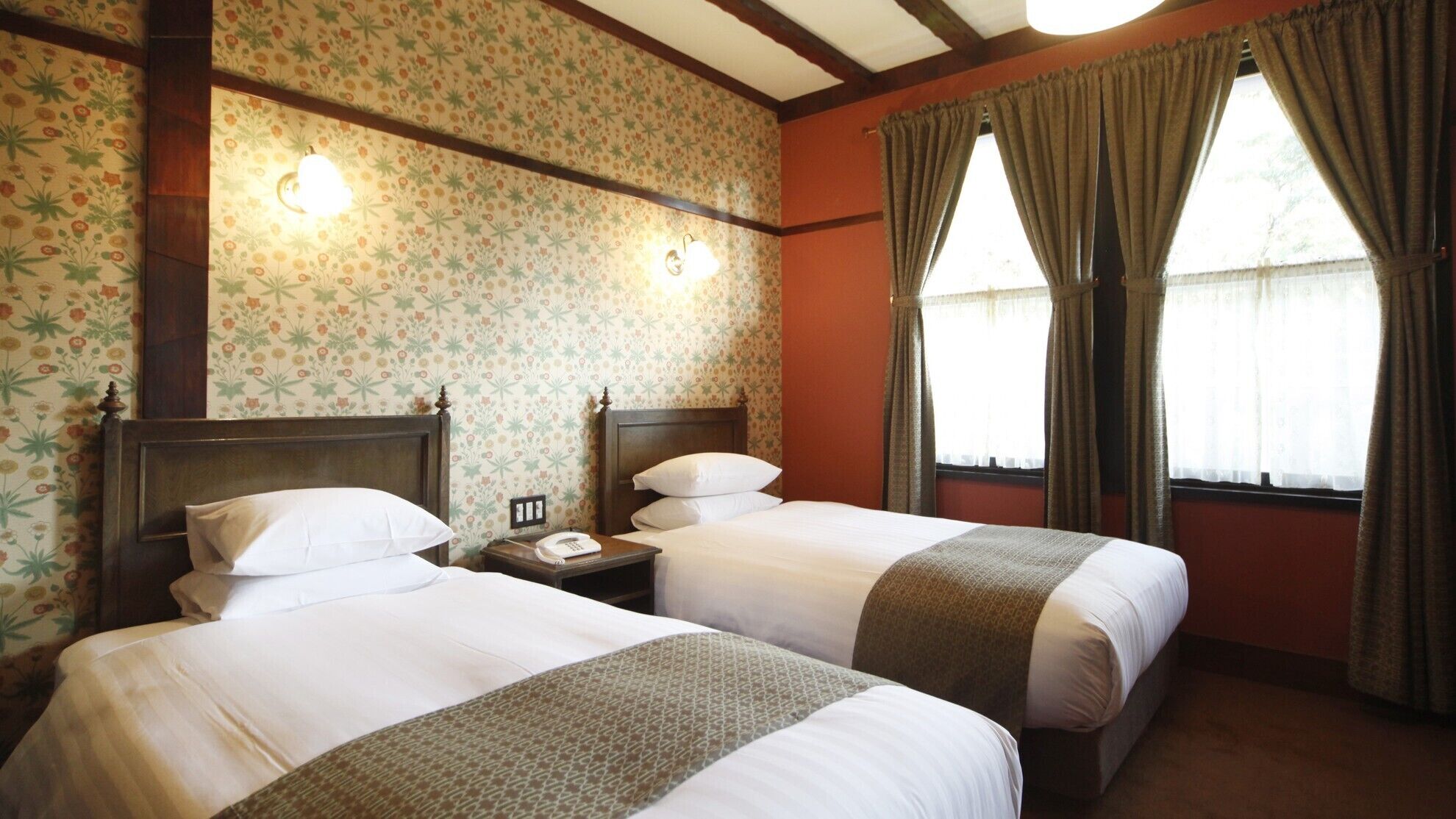 A hotel room with two beds, floral wallpaper, wooden beams, a nightstand with a phone, and a window with curtains.