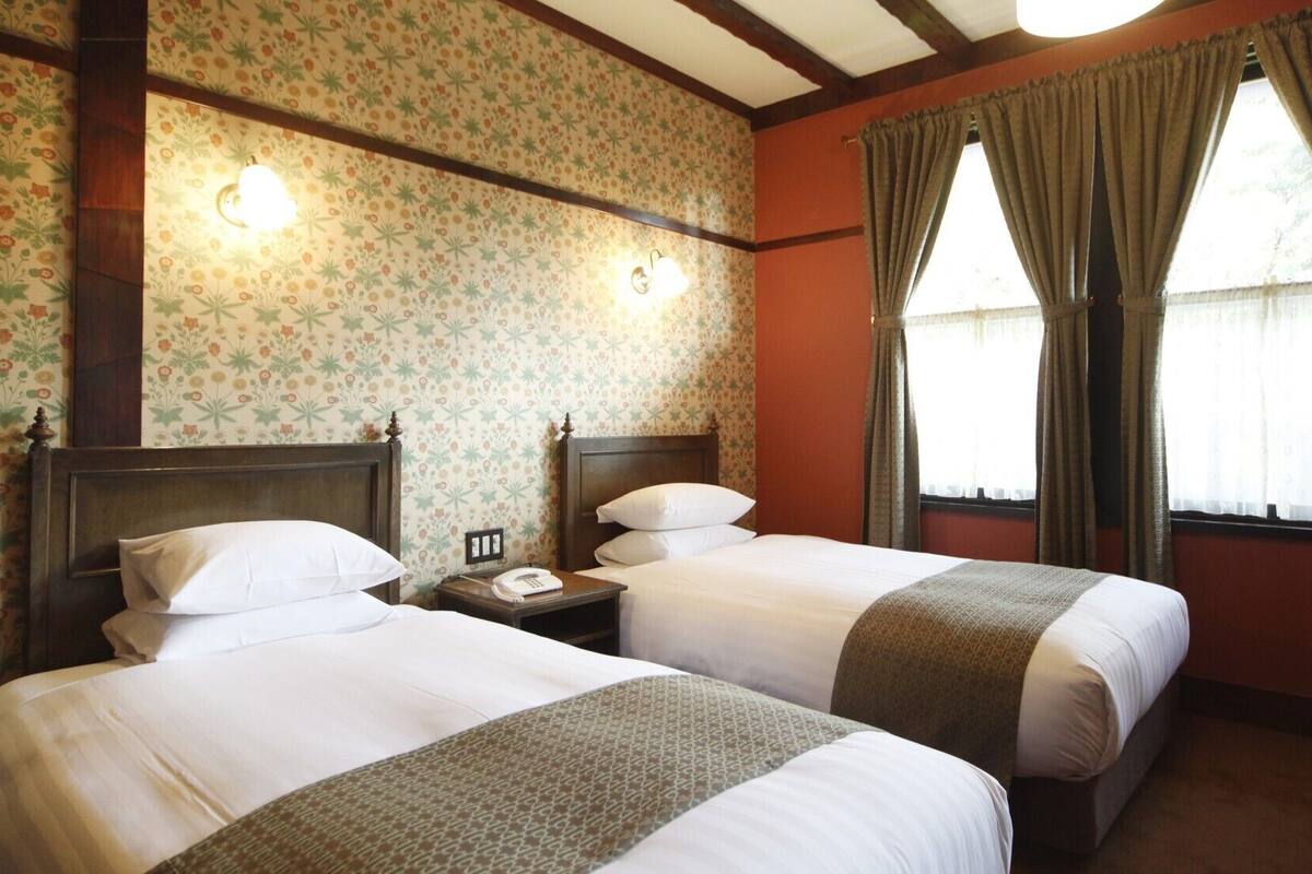 A hotel room with two beds, floral wallpaper, wooden beams, a nightstand with a phone, and a window with curtains.