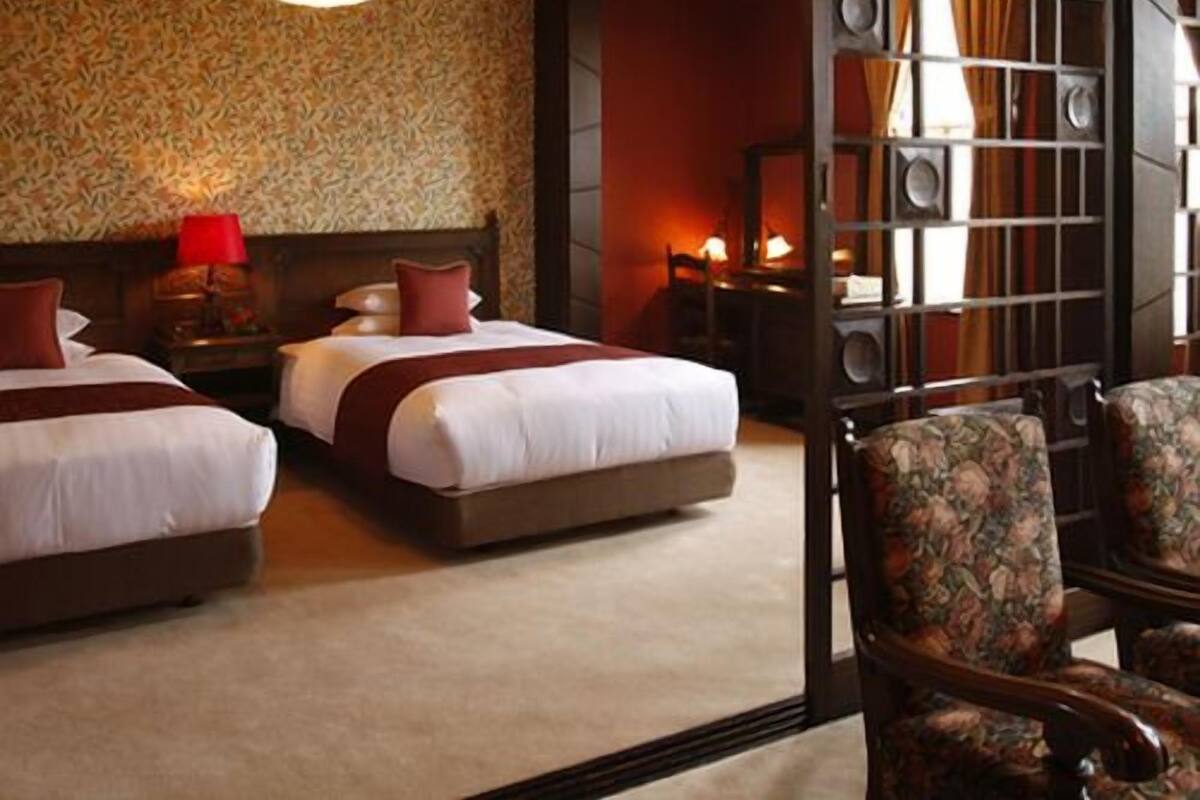 A hotel room with two beds, a wooden ceiling, a patterned wall, and a seating area with a chair.