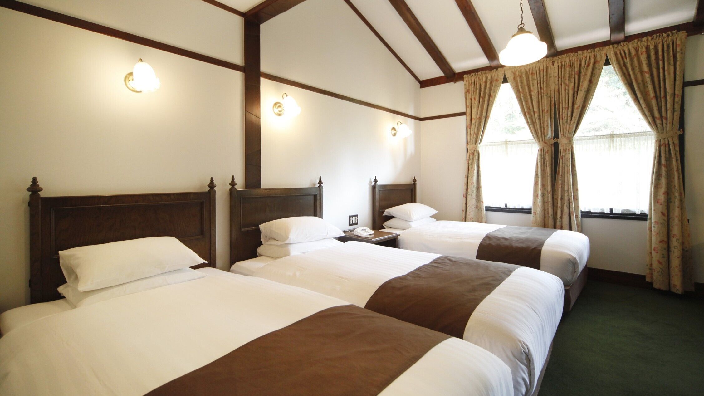 A hotel room with three single beds, wooden headboards, wall-mounted lights, a telephone, and a window with curtains.