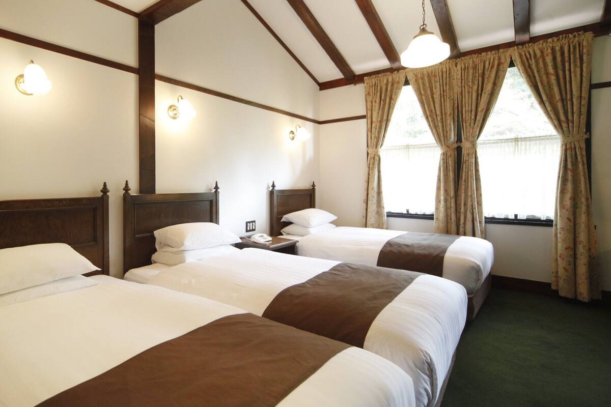 A hotel room with three single beds, wooden headboards, wall-mounted lights, a telephone, and a window with curtains.