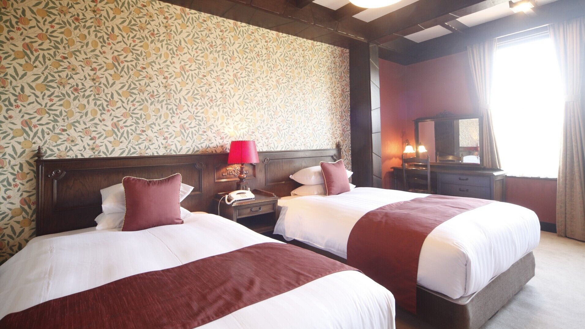 A hotel room with two beds, a wooden headboard, a telephone, a lamp, a mirror, and a window with curtains.