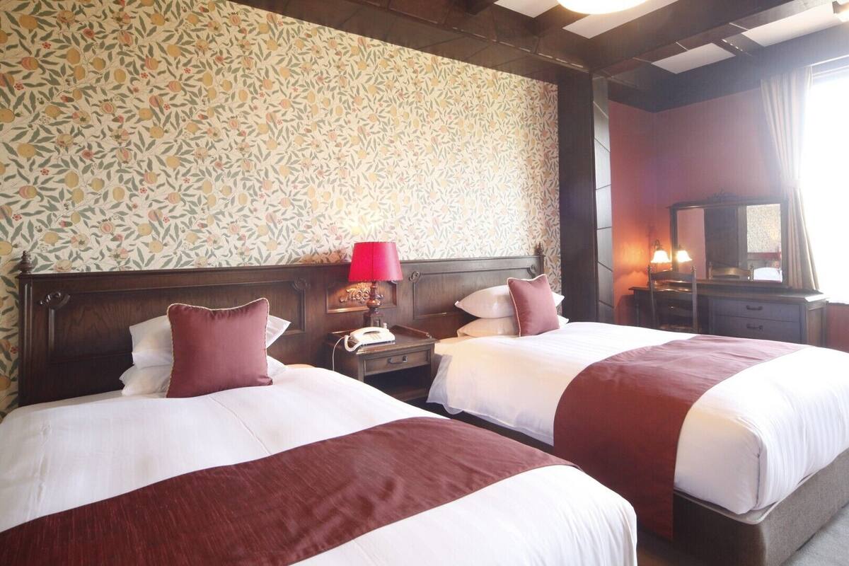 A hotel room with two beds, a wooden headboard, a telephone, a lamp, a mirror, and a window with curtains.