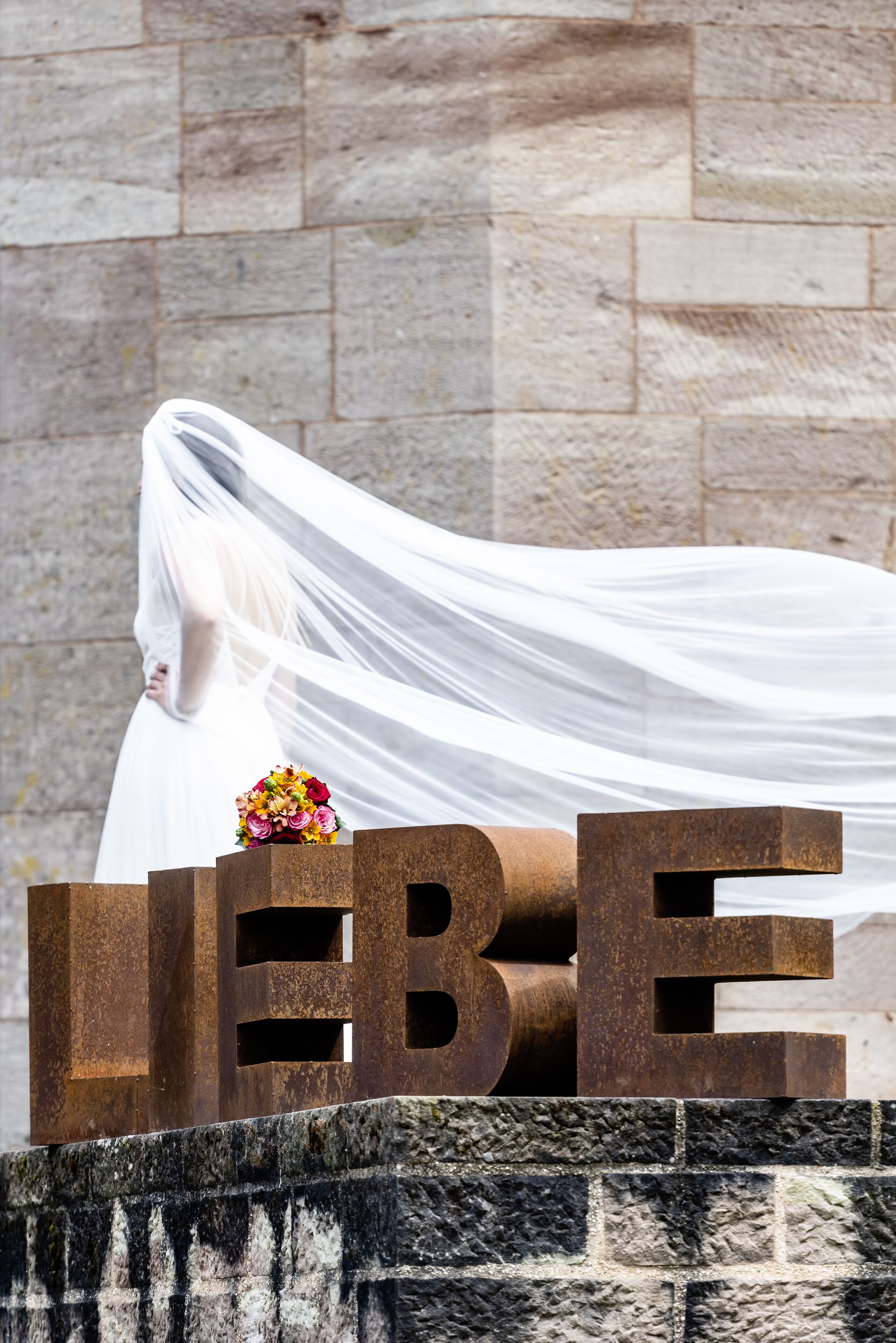 A stone wall with a metal structure featuring the word "LIEBE" and a bouquet of flowers.