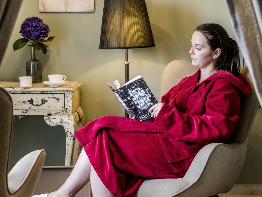 A person seated in a chair reading a book, with a lamp and a vase with flowers on a side table.