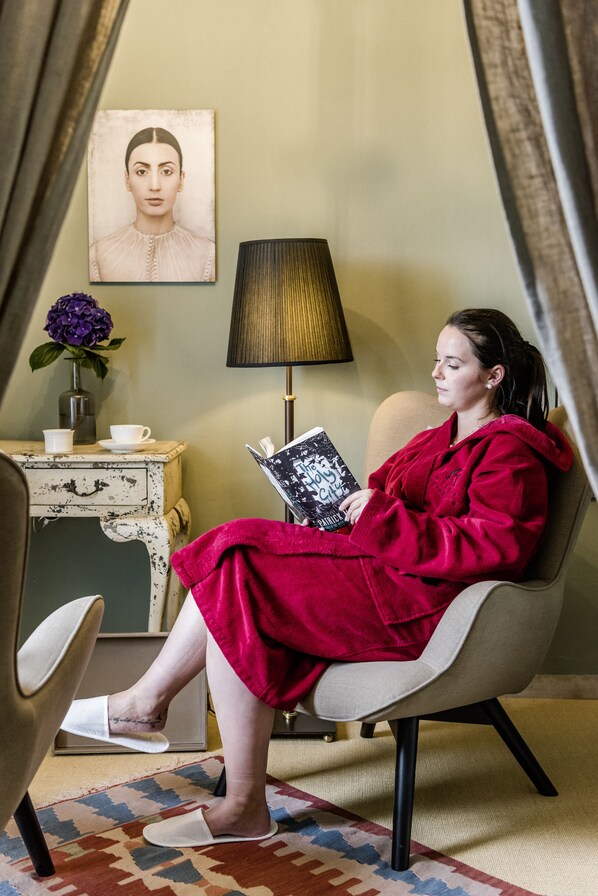 A person seated in a chair reading a book, with a lamp and a vase with flowers on a side table.