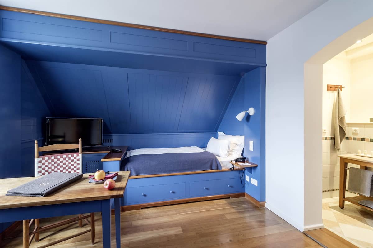 Un dormitorio con una pared azul decorativa, una cama con ropa blanca, un escritorio con una computadora portátil y una mesita de noche con una lámpara.