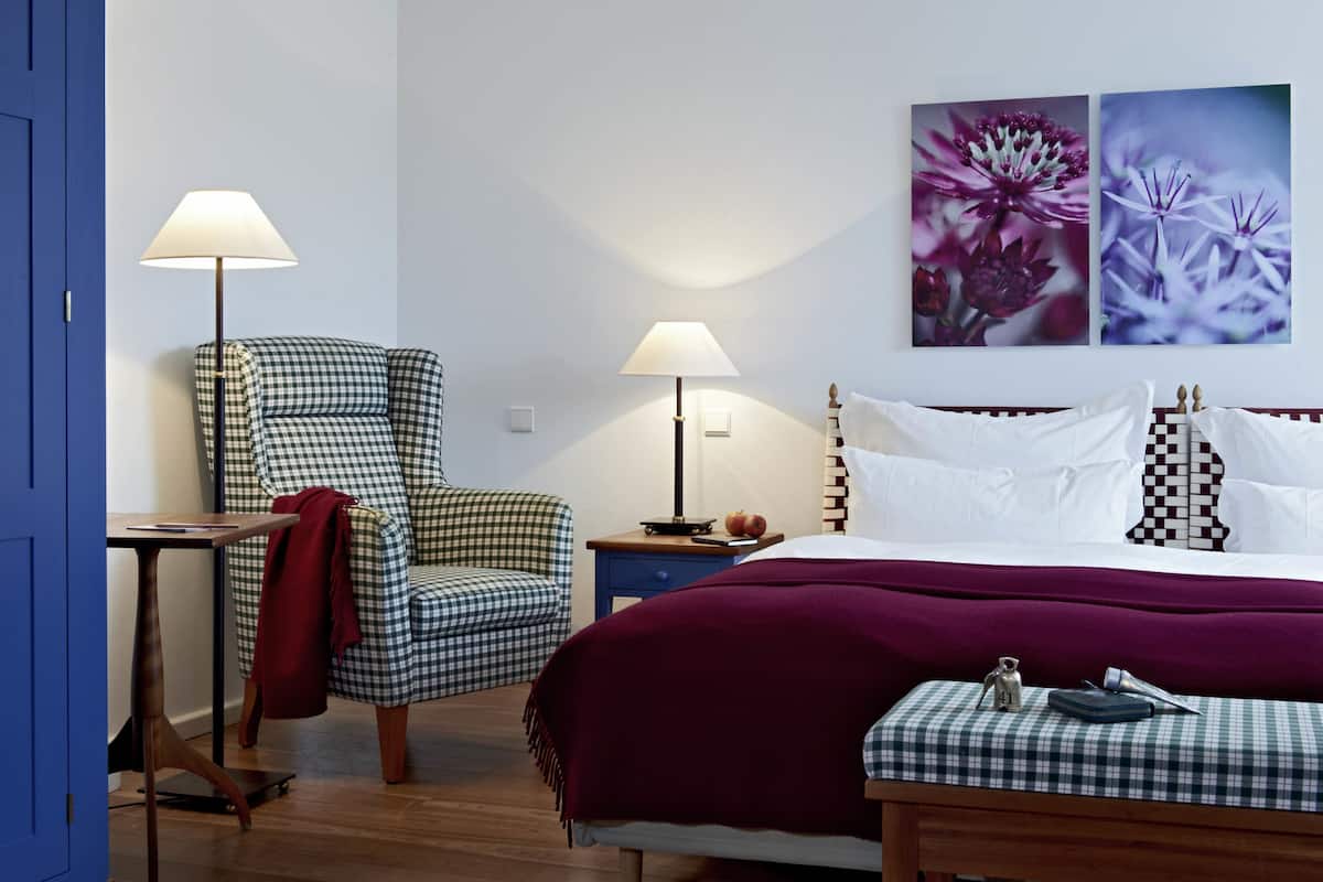 Un dormitorio ordenado con un sillón a cuadros, una cama con manta morada, dos lámparas de noche y un cuadro floral en la pared.