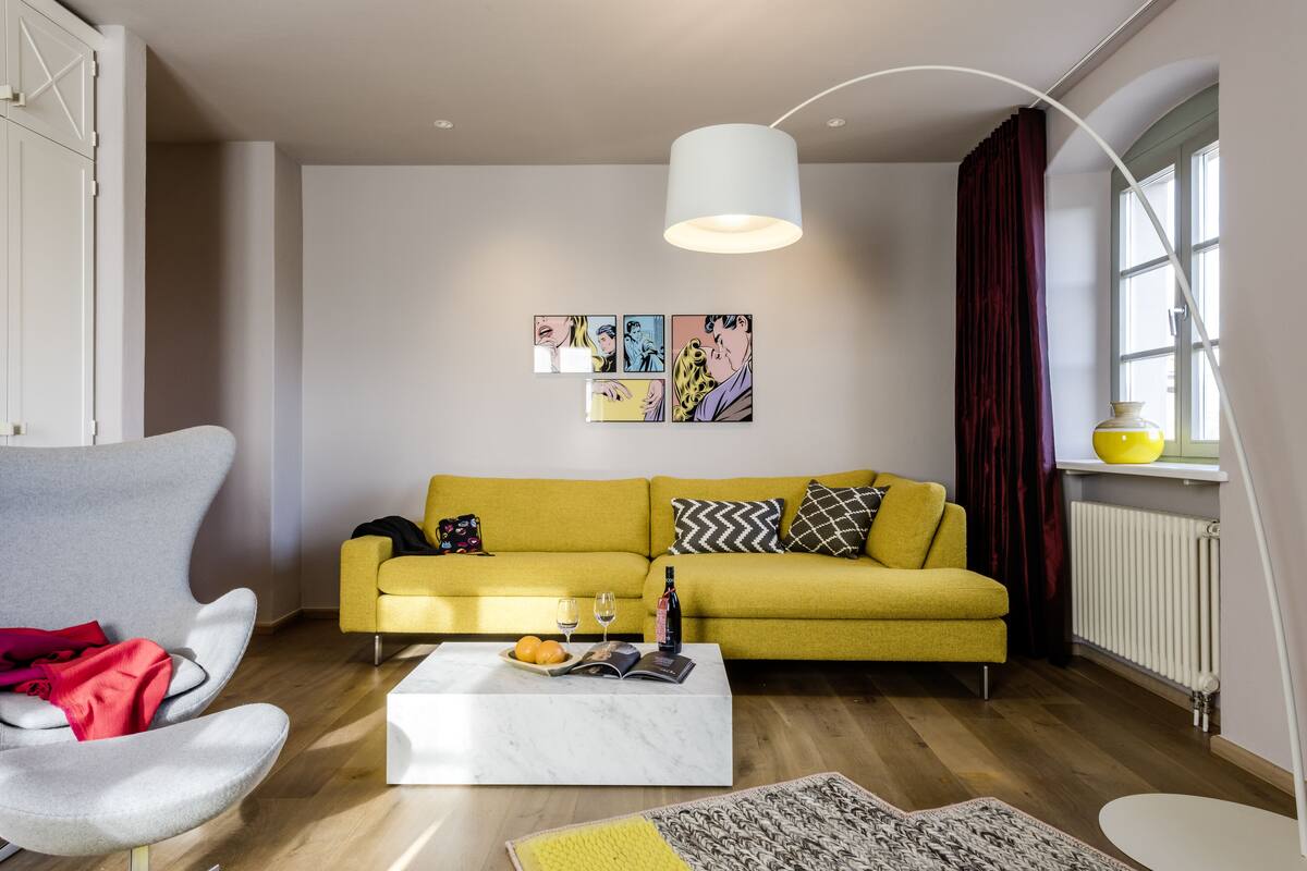 Una sala de estar moderna con un sofá amarillo, un sillón blanco, una mesa de centro de mármol y un ventanal con cortinas.