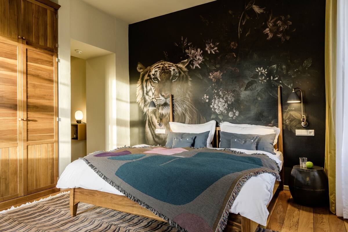Un dormitorio con una cama grande, un armario de madera, un mural en la pared, una lámpara de noche y una mesita con un vaso y una manzana.