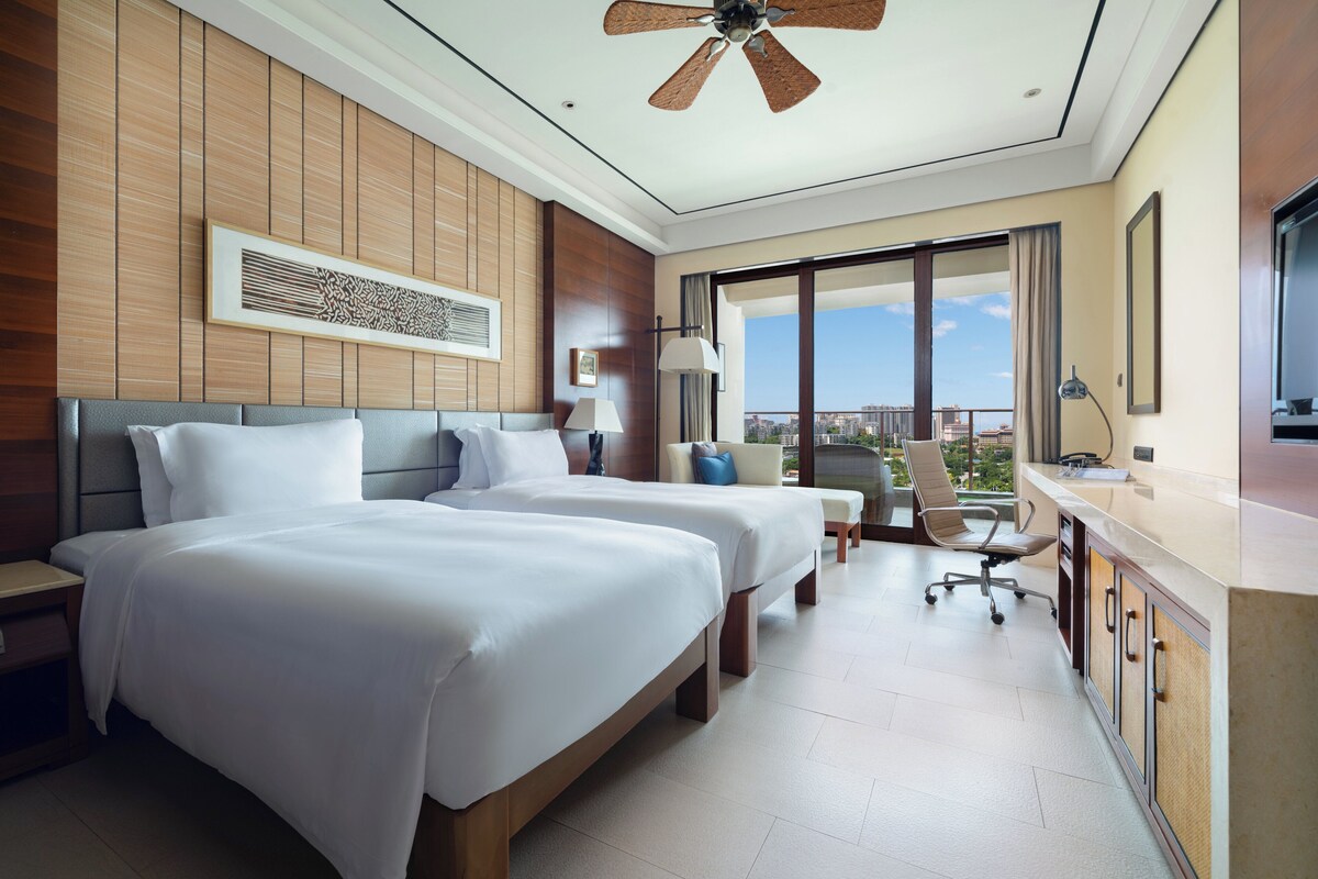 Ein Hotelzimmer mit zwei Betten, einem Schreibtisch, einem Stuhl, einem Deckenventilator und einem großen Fenster mit Blick auf die Stadt.