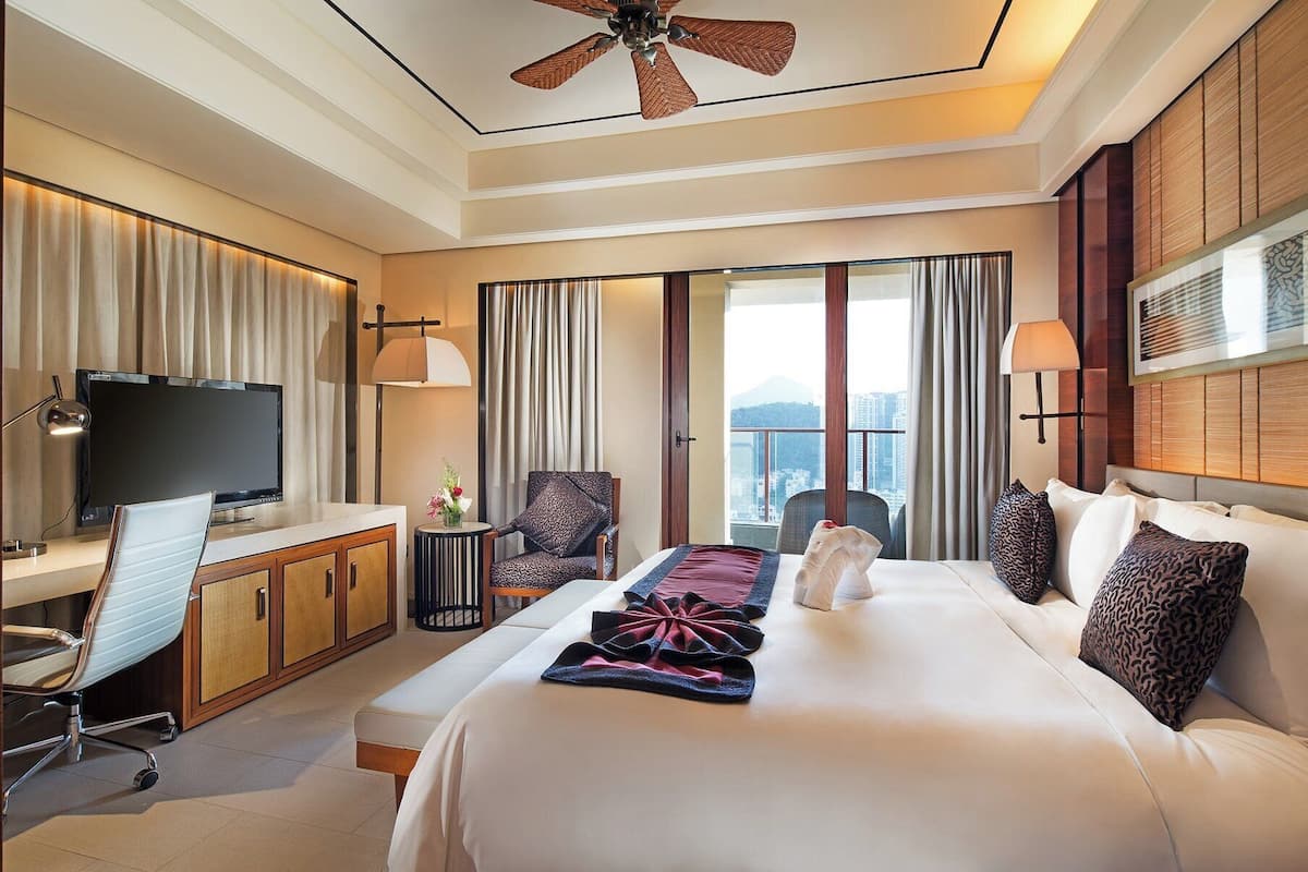 Habitación de hotel con una cama grande, un escritorio con televisor, una silla, una lámpara y vistas a la ciudad.