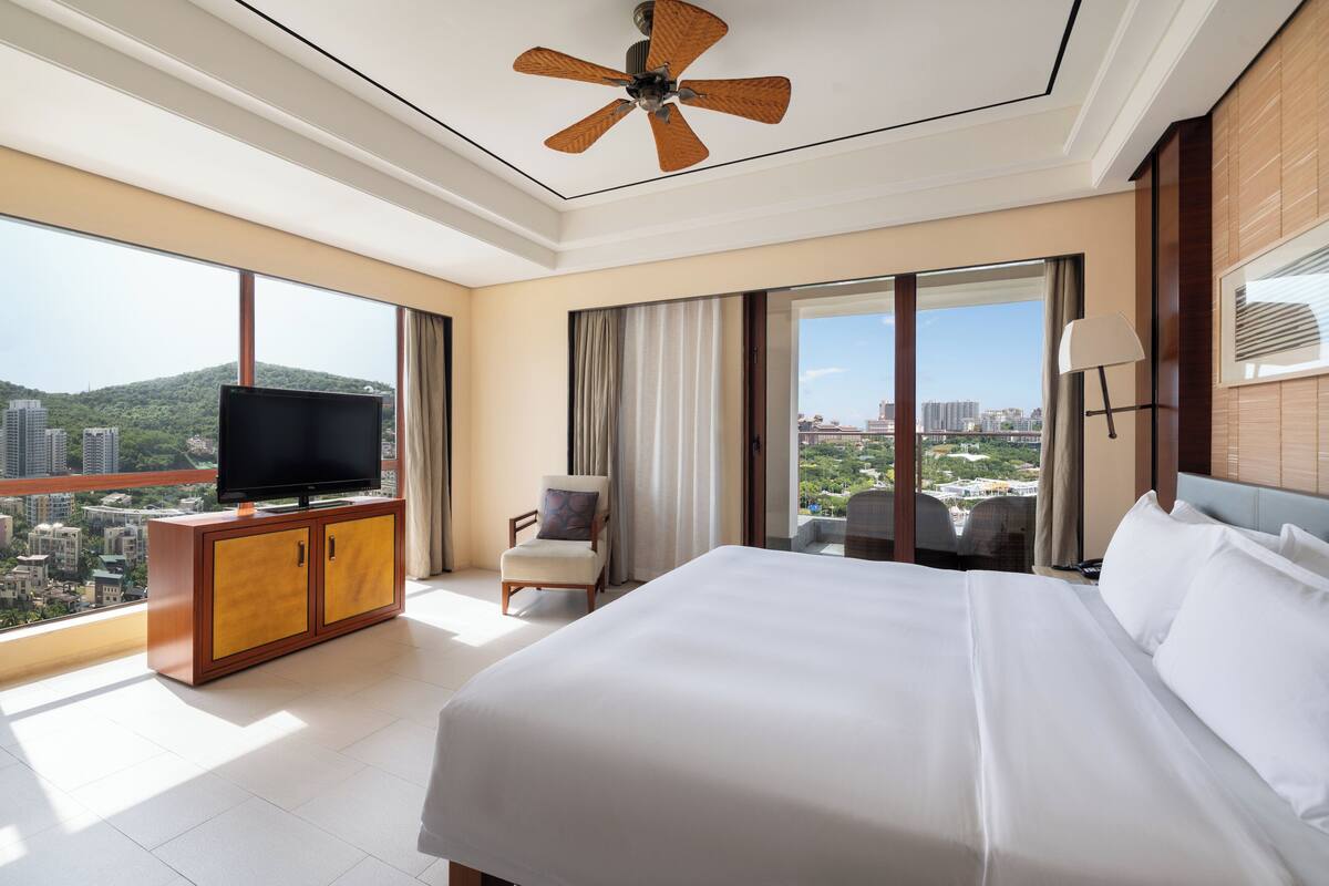 Ein Hotelzimmer mit einem großen Bett, einem Fernseher auf einer Holzkommode, einem Sessel und Blick auf die Stadt.