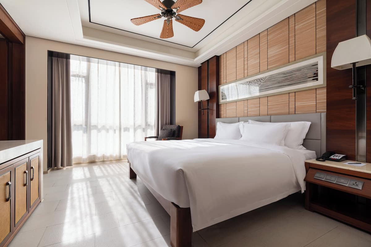 Habitación de hotel con una cama grande, un escritorio con lámpara, un ventilador de techo y un ventanal con cortinas.
