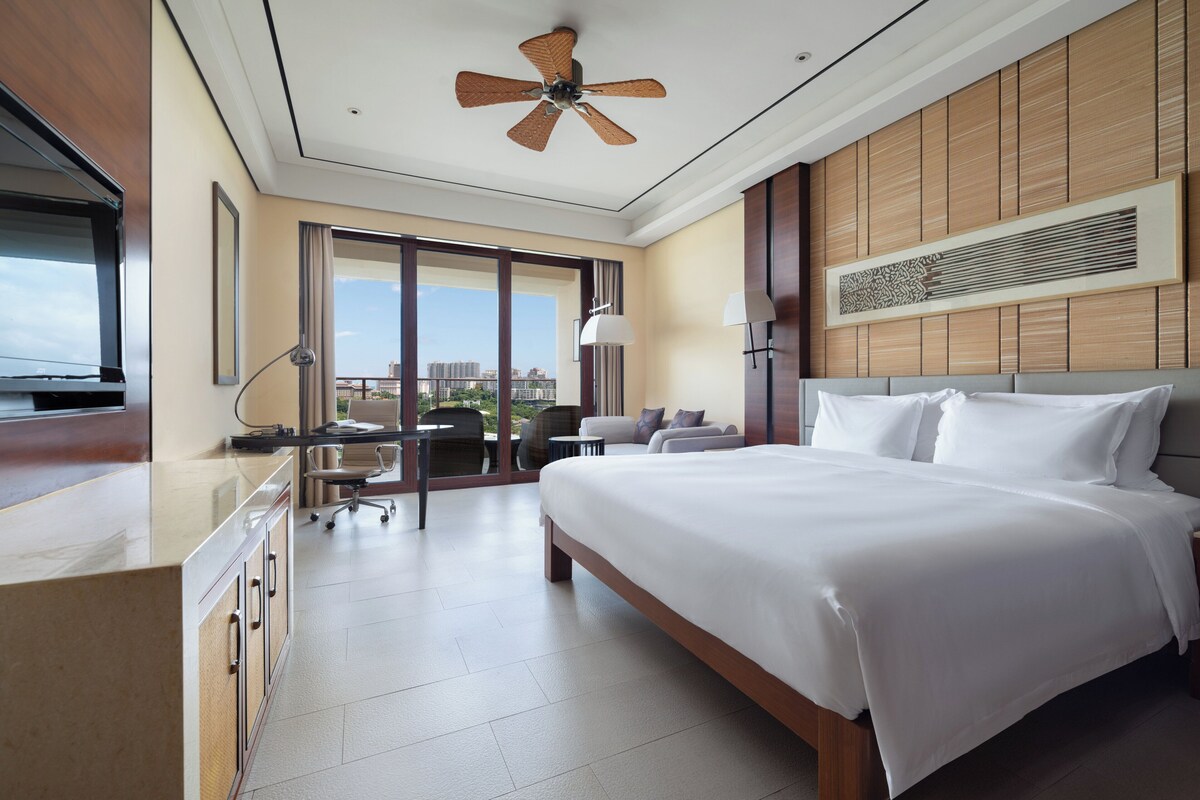 Ein Hotelzimmer mit einem großen Bett, einem Schreibtisch mit Stuhl, einem Deckenventilator und Blick auf die Stadt durch große Fenster.