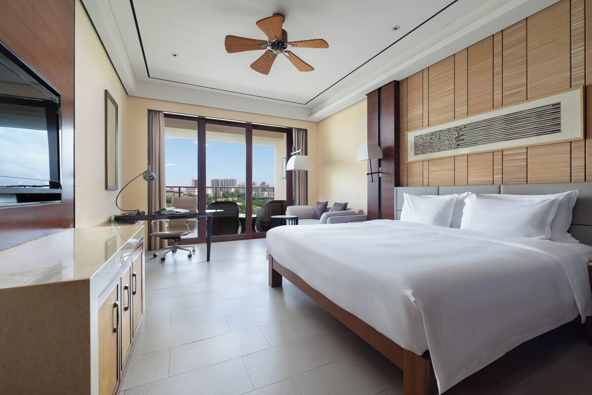 Ein Hotelzimmer mit einem großen Bett, einem Schreibtisch mit Stuhl, einem Deckenventilator und Blick auf die Stadt durch große Fenster.
