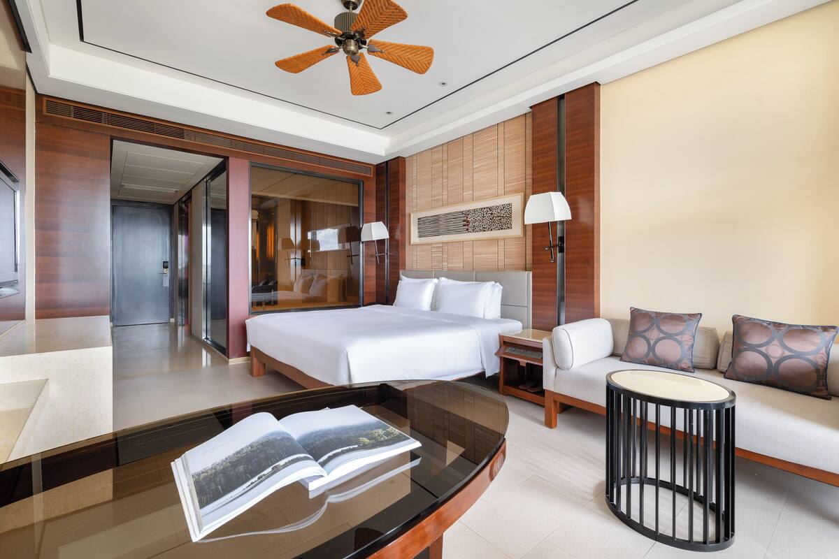 Una habitación de hotel moderna con una cama grande, un sofá, una mesa de centro de vidrio y un ventilador de techo.