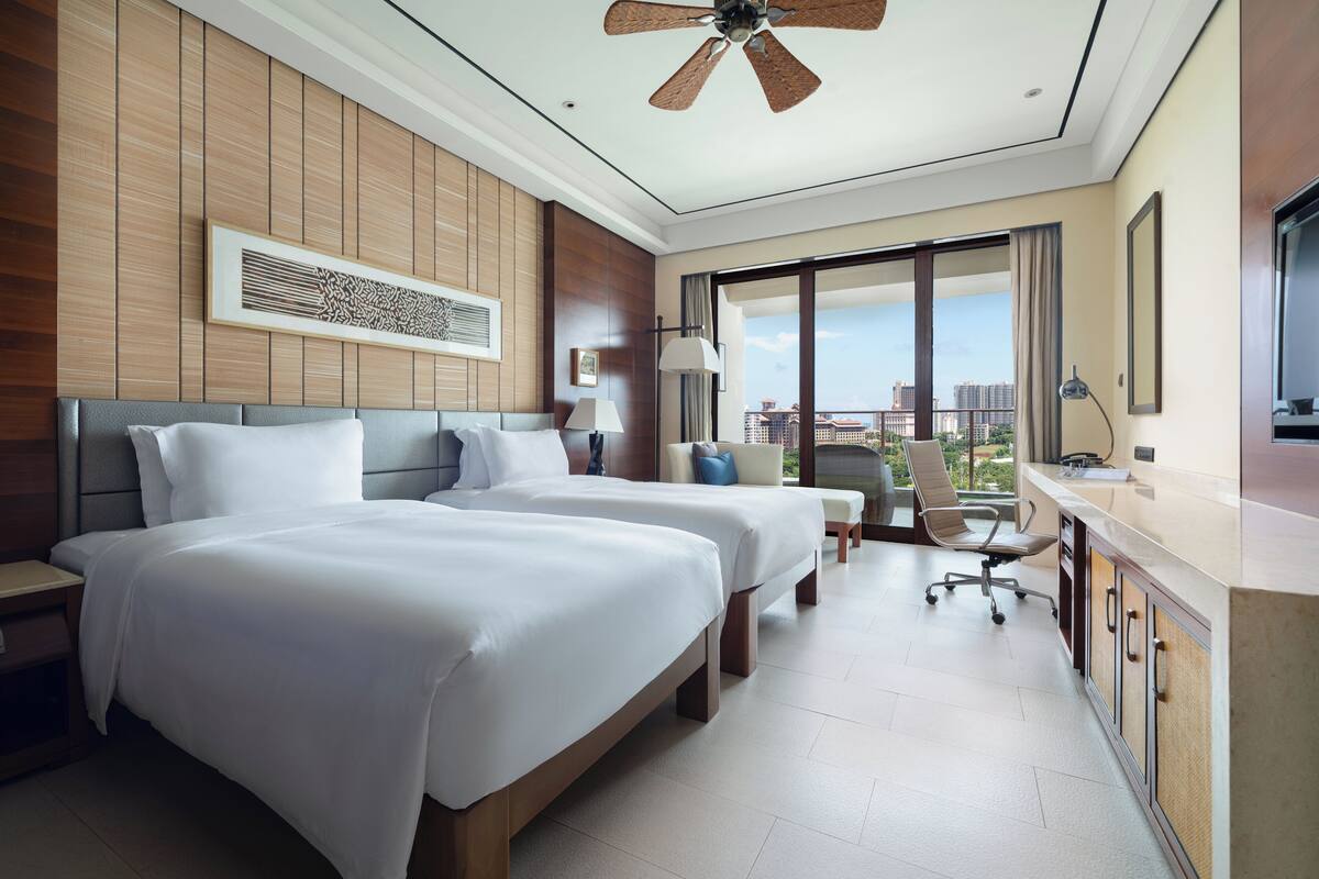 Habitación de hotel con dos camas, un escritorio, una silla, un ventilador de techo y un amplio ventanal con vista a la ciudad.