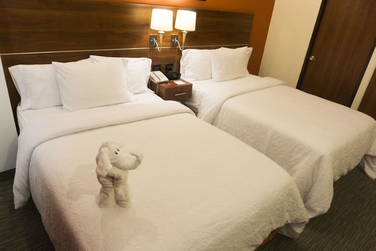 A hotel room with two beds, a nightstand with a phone, two wall-mounted lamps, and a teddy bear on the bed.
