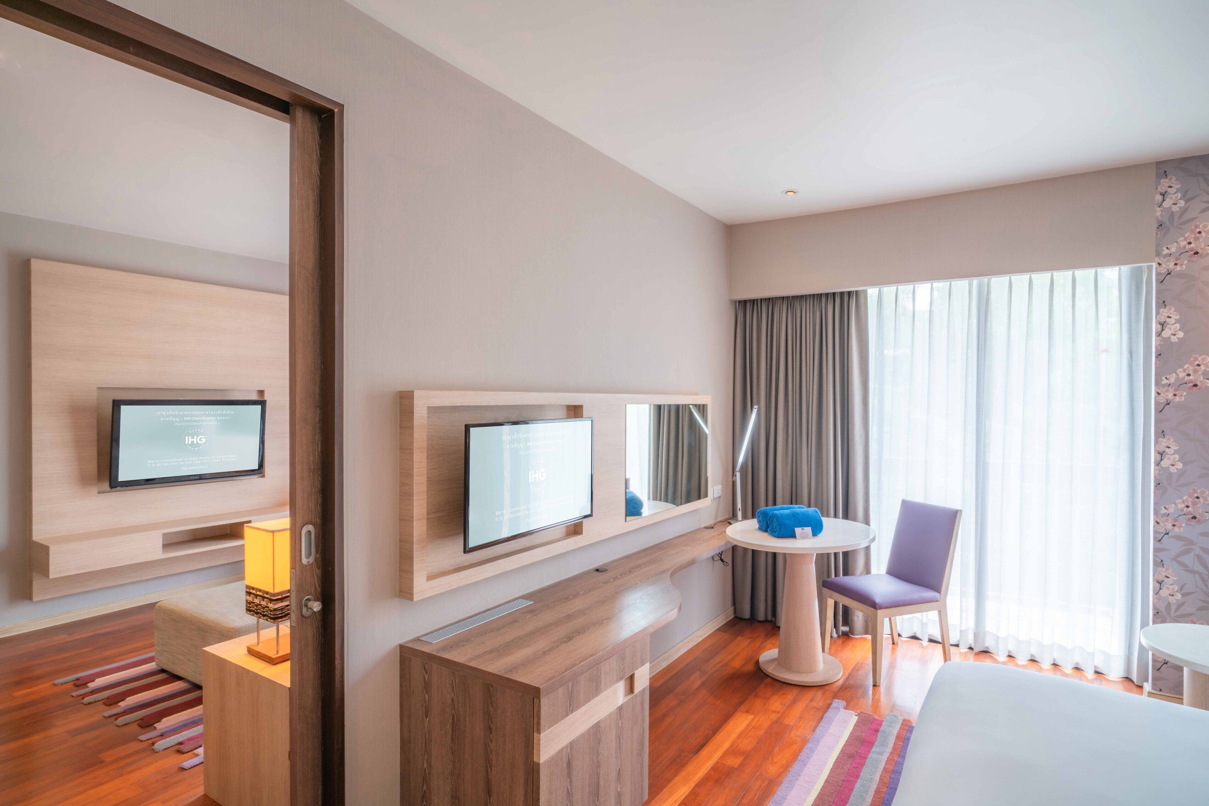 A modern hotel room with a wooden floor, a bed, a desk with a computer, a TV, a small table with a chair, and a large window with curtains.