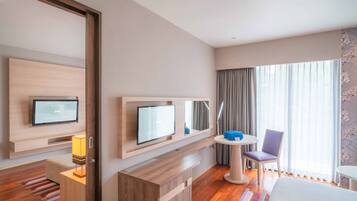 A modern hotel room with a wooden floor, a bed, a desk with a computer, a TV, a small table with a chair, and a large window with curtains.
