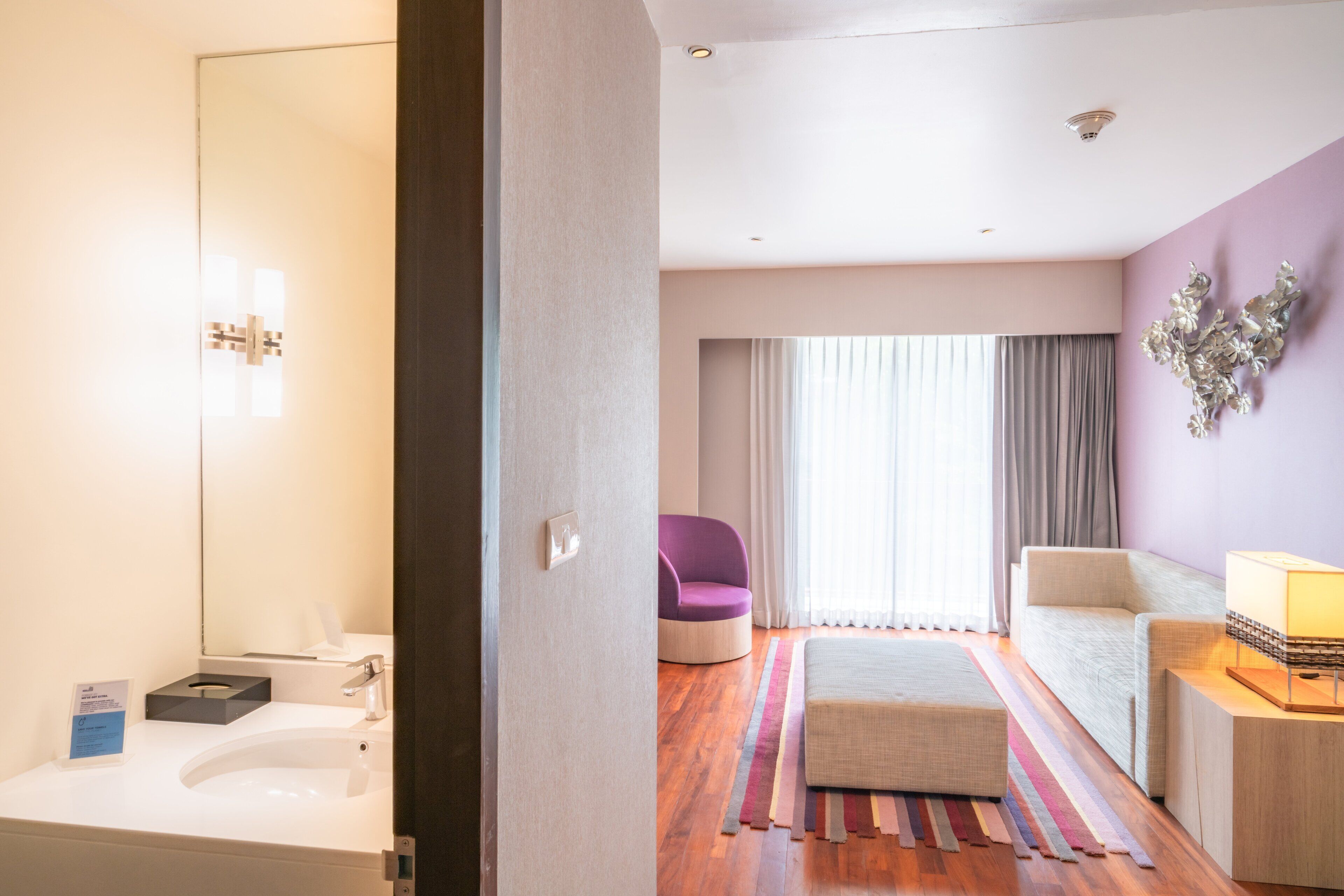 A modern hotel room with a white sink, a purple chair, a beige sofa, and a striped rug.