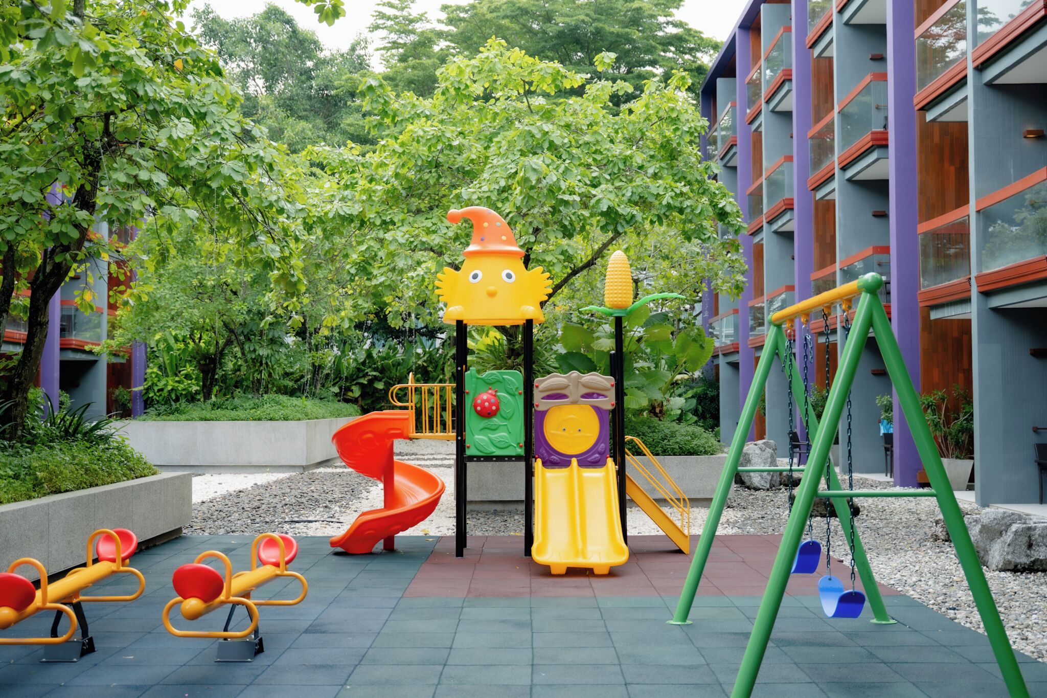 A playground with various play structures, including a slide, swings, and seesaws, surrounded by greenery and a building with large windows.
