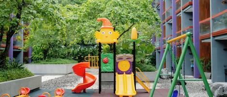 A playground with various play structures, including a slide, swings, and seesaws, surrounded by greenery and a building with large windows.