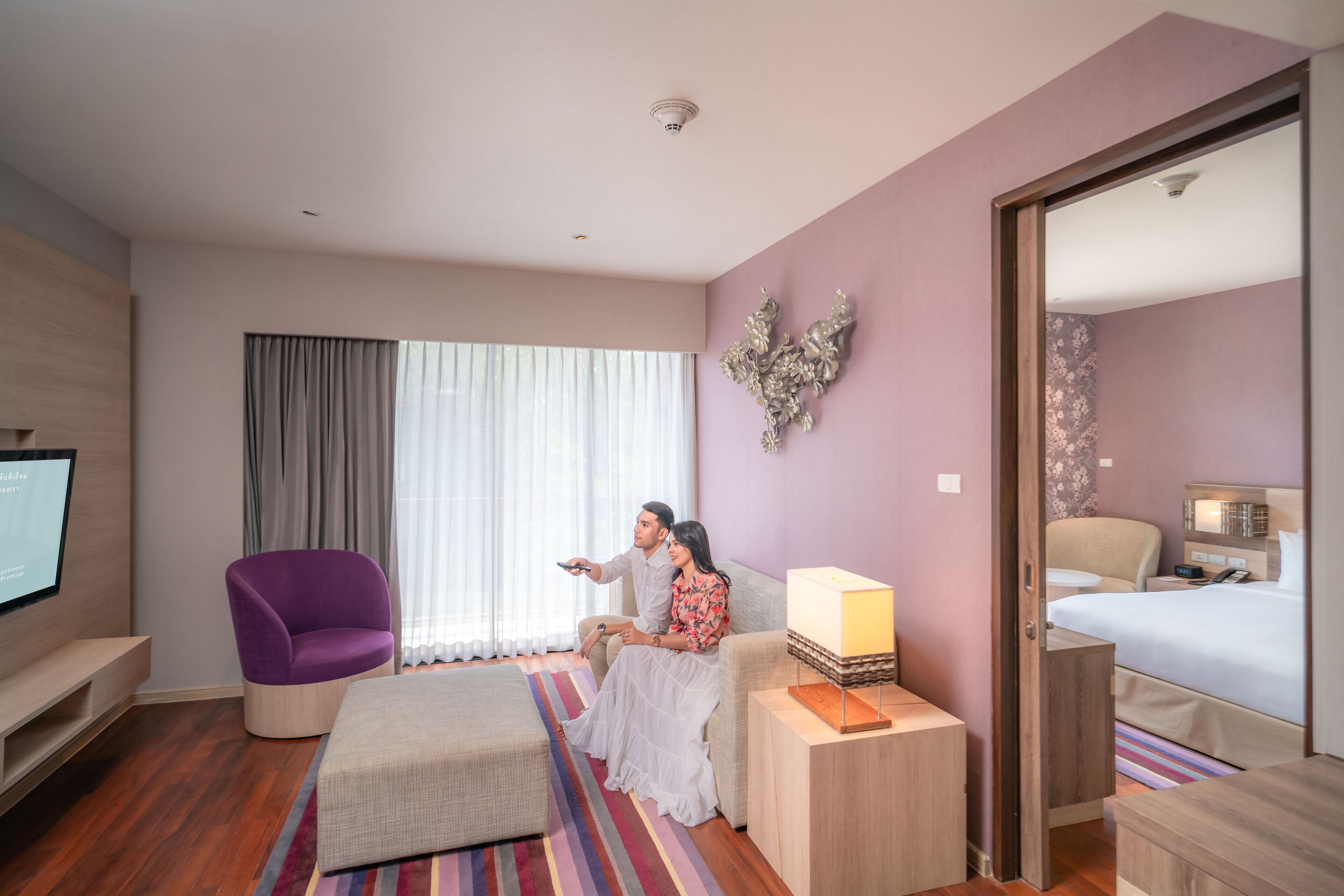 A hotel room with a large bed, a TV, a purple armchair, and a small table with a lamp.