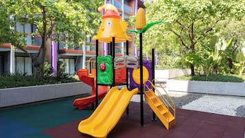 A playground with slides and climbing structures, surrounded by trees and a building in the background.